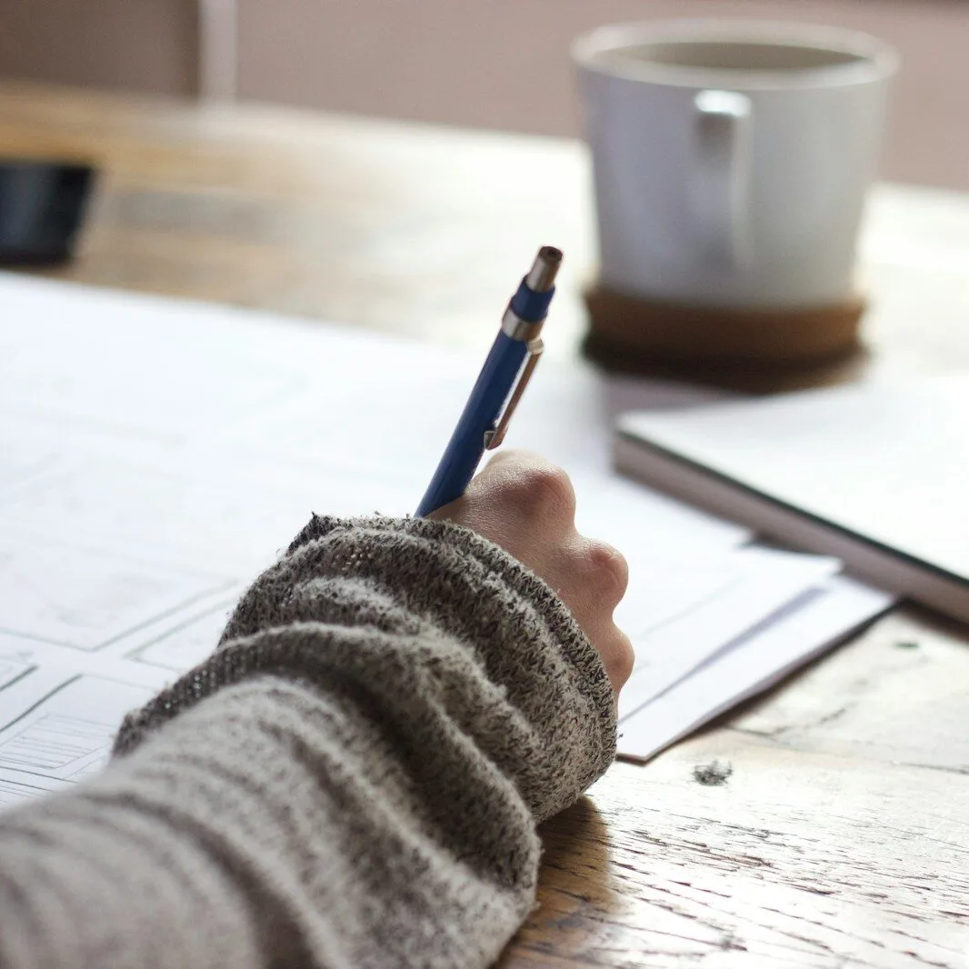 person writing on brown wooden table near white ceramic mug
